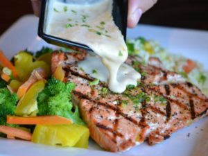 Person pouring sauce onto grilled salmon with vegetables
