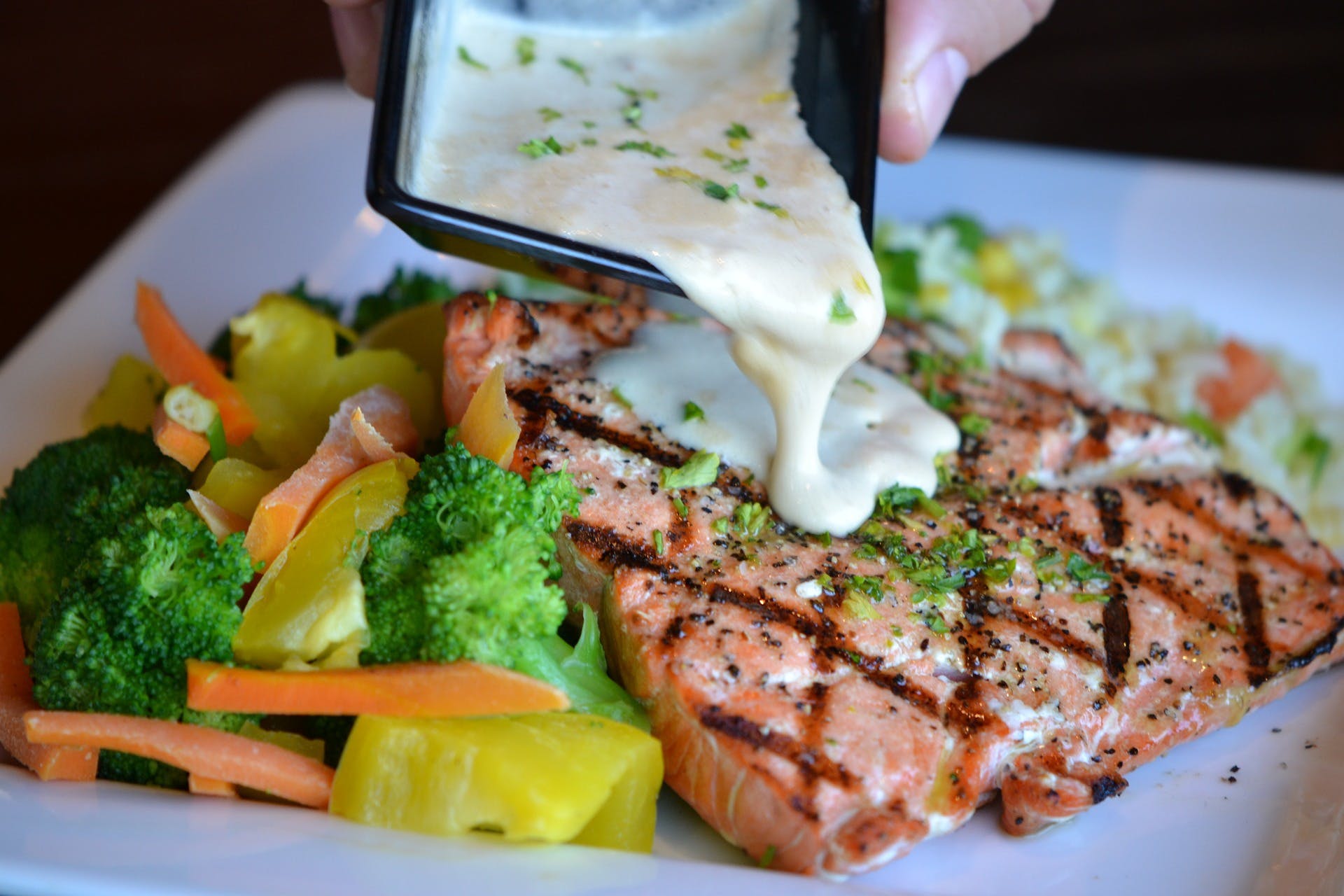 Person pouring sauce onto grilled salmon with vegetables